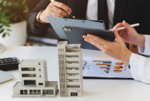 Two colleagues at a table reviewing printed copies of graph data and pointing out data on a tablet. Miniature of building structure is on the table.