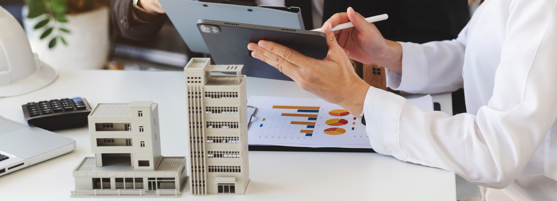 Two colleagues at a table reviewing printed copies of graph data and pointing out data on a tablet. Miniature of building structure is on the table.