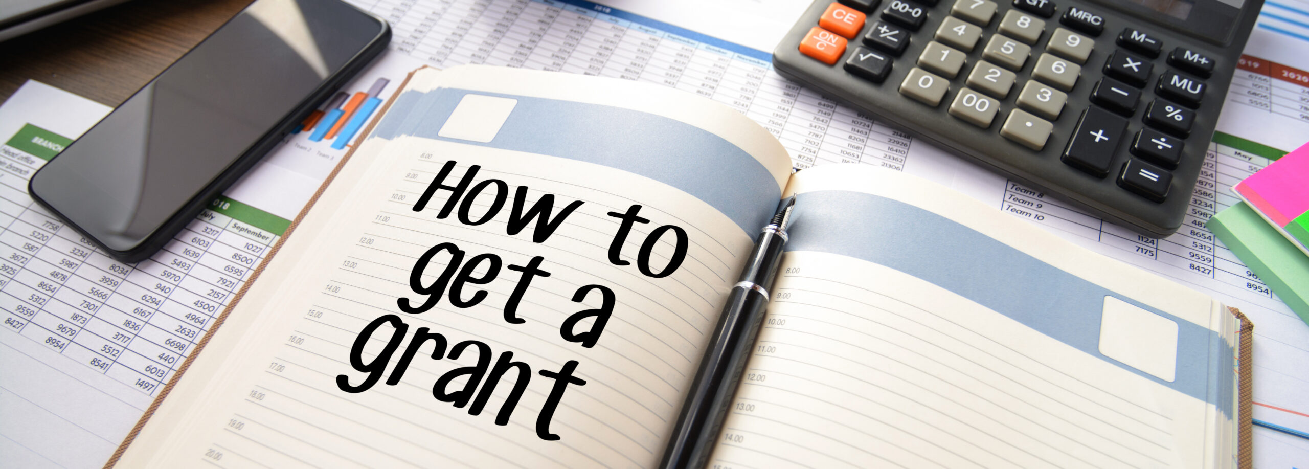 Business accessories on a table, including a smartphone, calculator, reports and notepad with text "How to get a grant"