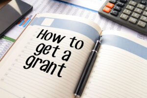 Business accessories on a table, including a smartphone, calculator, reports and notepad with text "How to get a grant"