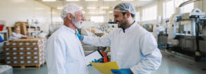 Two men in hair nets and white lab coats smiling at each other in food processing plant