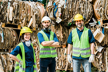 Three recycling center employees in hard hats and safety vests in front of bundled cardboard