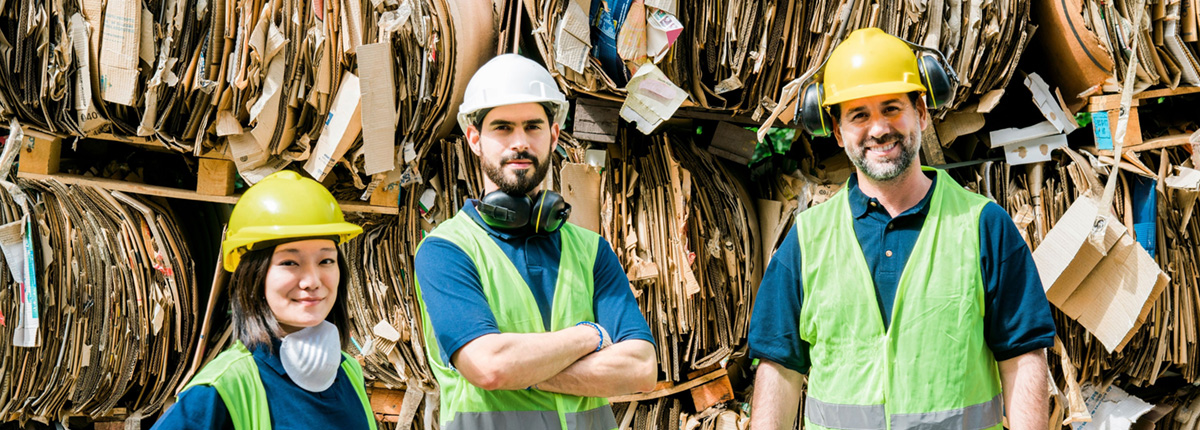 Three recycling center employees in hard hats and safety vests in front of bundled cardboard