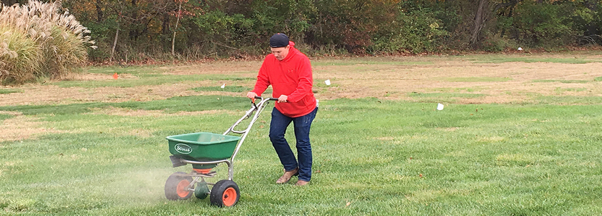 Man in red shirt pushing pesticide spreader over grass field