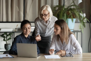 Mature businesswoman mentor in glasses training two younger women, she is pointing at laptop screen