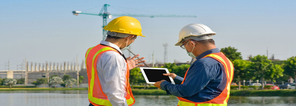 two men in hard hats and vests standing outside looking at tablet with crane in background