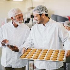 <a href="/food-science-safety">Food Science & Safety</a> Two employees in sterile clothes in food factory smiling and talking, one holding tablet and one holding tray of cookies
