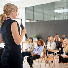 <a href="/employee-training">Customized Staff Training</a> Woman holding microphone speaking to room of professionals