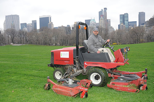 Central Park Conservancy staff on ride-on mower with NYC skyline in the background