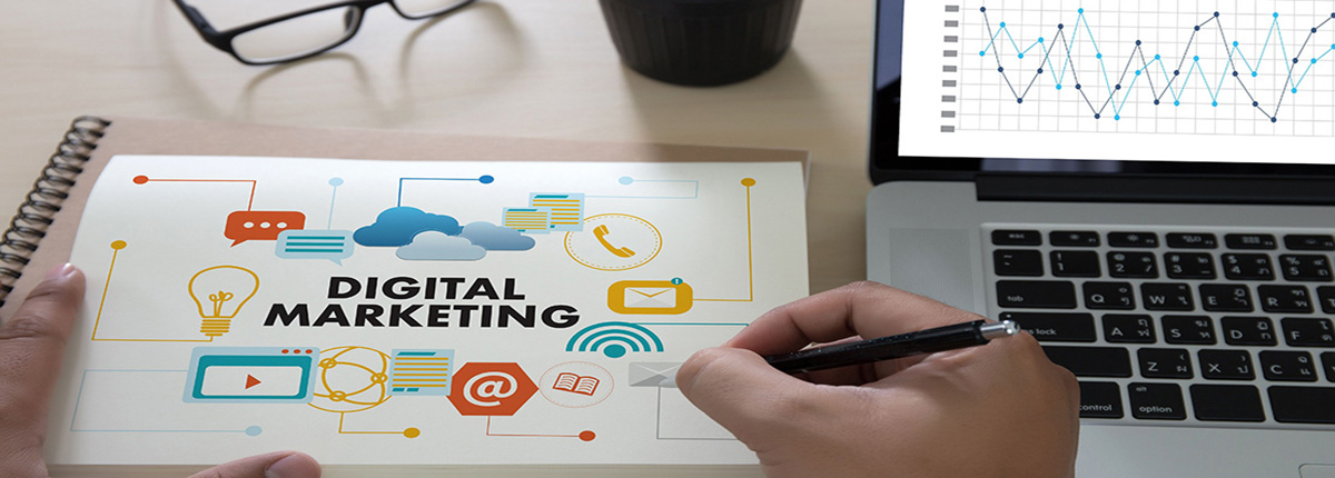 Desk scene with laptop, pen, and hand holding pen above notebook reading Digital Marketing