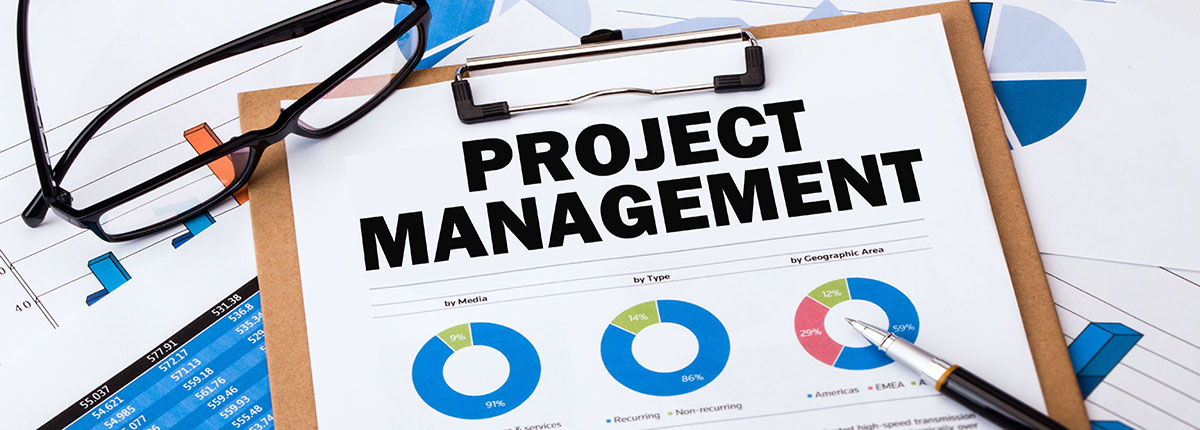 Clipboard with project management sheet on it surrounded by pair of glasses, pen, and papers with charts on them