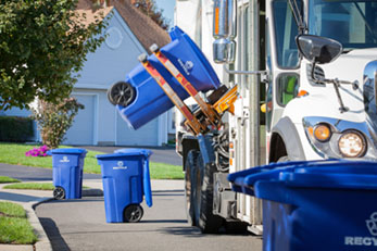 Mechanical arm of recycling truck picking up blue recycling can on residential street