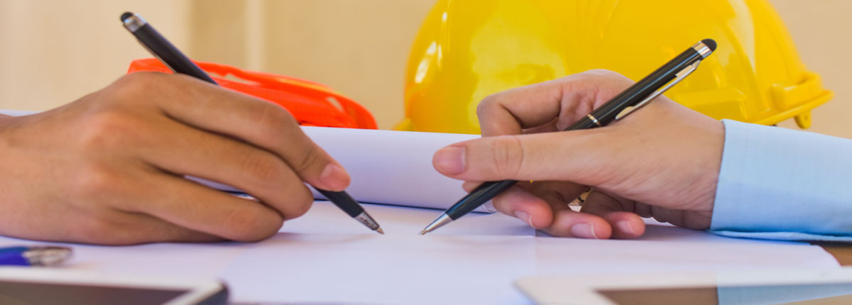 Closeup of two people's Hands Writing with hardhat in the background and tablets in the foreground